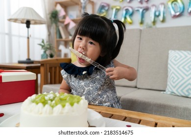 Closeup Of Naughty Asian Baby Girl Sneaking A Taste And Licking Yummy Cream On Cake Knife While Cutting Cake At Birthday Party At Home