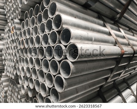 A close-up of a neatly stacked bundle of galvanized metal pipes, creating a pattern of circular openings and reflective surfaces. The image highlights industrial strength and precision.