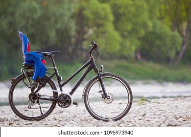 Close-up of new modern black bicycle with blue plastic child seat standing on side stand on lit by sun pebbles on blurred green trees summer bokeh background. Car free day concept.
