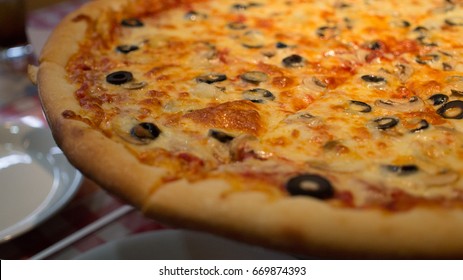 Closeup Of A New York Style Pizza With Extra Cheese, Olives, And Tomato Sauce, Half In Frame With A White Plate In The Background