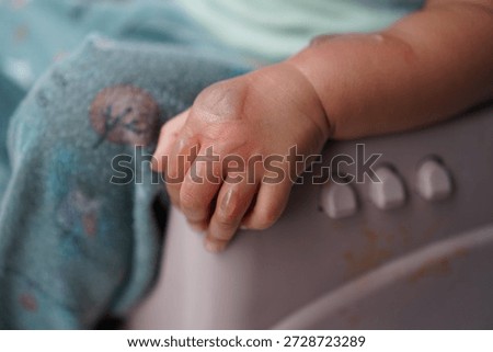 A close-up of a newborn's tiny hand gently gripping the edge of a baby seat with soft fabric and rounded buttons in the background conveying new life and tender care during early infancy.