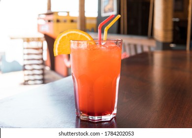 Closeup Ocktail Of Sweet Bourbon Whiskey Drink Garnished By Orange Peel On Table Diner Booth With Blurry Bokeh Restaurant Background. A Glass Of Red Juice On A Wooden Table