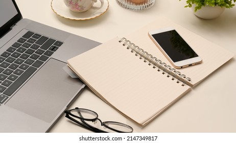 Closeup, Office Desk Workspace With Open Spiral Notebook, Smartphone, Eyeglasses And Laptop On Table