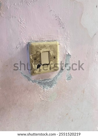 A close-up of an old and damaged light switch on a wall. The switch plate is discolored and cracked, showing visible signs of wear and tear. The surrounding wall has peeling paint and moisture damage.