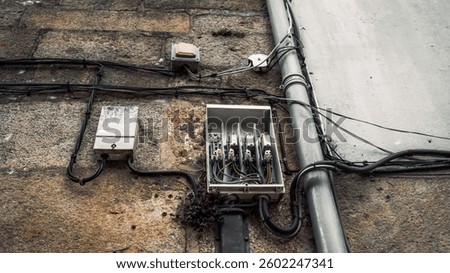 Close-up of an old electrical distribution box with open cover and exposed wiring mounted on a weathered stone wall, along with cables and conduit pipes, outdoors during daytime