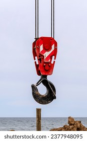 Closeup of an old lifting hook attached to sheave suspended by wire rope from boom of crane against clear blue sky. Construction work on the seashore