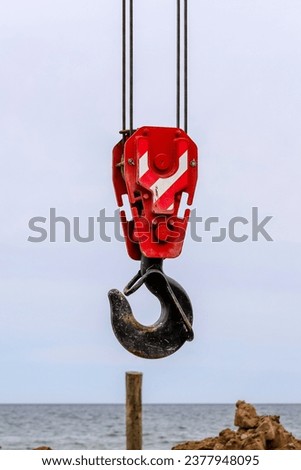 Closeup of an old lifting hook attached to sheave suspended by wire rope from boom of crane against clear blue sky. Construction work on the seashore
