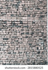 Close-up of an old rustic brick wall with weathered beige and grayish-green bricks. The bricks have uneven texture, patches of wear, and cement mortar between them. Some bricks are darker and some are lighter, giving the wall a vintage, aged look. The wall shows natural imperfections and a horizontal band of larger bricks in the middle.
