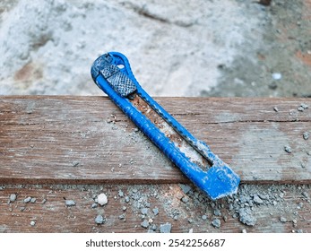 Close-up of an old, rusty blue utility knife placed on wooden surface with dust and small debris around, giving a rugged, worn-out appearance. Construction tool, and industrial environments. - Powered by Shutterstock