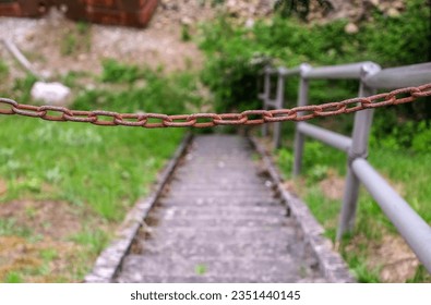 Close-up of an old, rusty chain in front of a stone staircase with a metal railing and some overgrown vegetation, selective focus - Shutterstock ID 2351440145