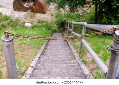 Close-up of an old, rusty chain in front of a stone staircase to s site, with a metal railing and some overgrown vegetation, selective focus - Shutterstock ID 2351446499