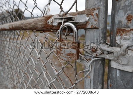 A close-up of an old, weathered padlock attached to a rusty metal latch on a chain-link fence gate, indicating security or closure.