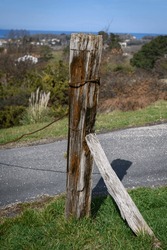 Close-up Of An Old Wooden Post Marked By The Passage Of Time From Which A Rusty Iron Cable Hangs, Part Of A Fence Another Smaller Pole Acting As A Support, In A Grassy Field At The Edge Of A Road