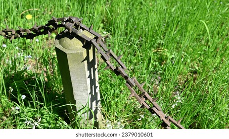 Close-up of an old wrought iron chain around a grave site consisting of rectangular chain links with thorn tips in sunshine and green grass in a cemetery in Germany