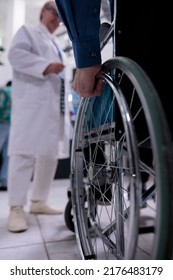 Closeup Of Older Man Hand Using Wheelchair In Front Of Medical Doctor For Appointment In Private Hospital Reception. Selective Focus On Senior Man Living With Disability At Medical Clinic.