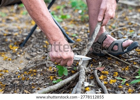 Close-up of an older white man using a knife to cut branches and twigs on the forest floor while wearing sandals outdoors.