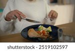 Close-up of an older woman cutting food on a plate, enjoying a nutritious meal with avocado, carrots, and chicken, emphasizing healthy eating habits and nutrition for seniors