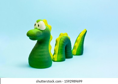 Close-up On Blue Background Of Decorative Toy Characterized As Loch Ness Monster, Pretending To Have Part Of His Body Submerged On The Support On Which It Rests.