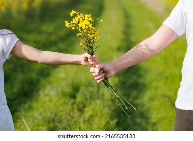 Close-up on bright small yellow bouquet with field wild small rapeseed flowers in hands of unknown teenage children, against backdrop of endless fields and green meadows under warm summer kind sun