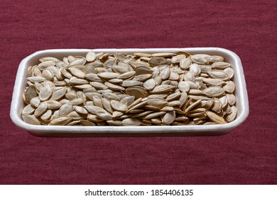 Close-up On A Burgundy Fabric Background, A White Disposable Container With Pumpkin Seeds.