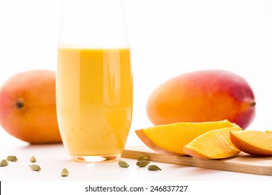 Close-up On Cut Mango Pieces On A Wooden Cutting Board. A Glass Filled With Mango Lassi, Cardamom Seeds And Two Entire Mangos. Selective Focus. Low Angle Shot. White Background And Bright Table Top.