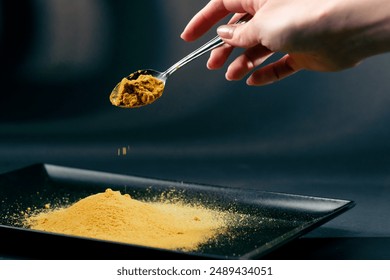 close-up on dark blue background with side lighting in a woman's hand, a spoon with the Indian spice turmeric is poured in doses into a black plate - Powered by Shutterstock