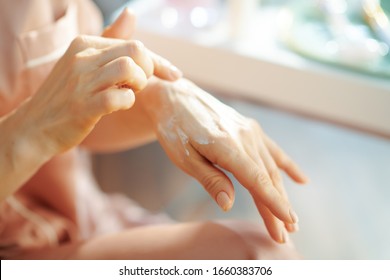 Closeup On Female In Pajamas Applying Hand Cream Near Table With Toiletries In The House In Sunny Winter Day.