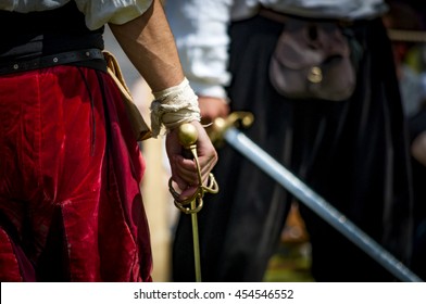 Closeup on the hand holding a sword of a pirate or medieval warrior, preparing to duel another man, there is a bandage on his wrist and the man in the background is blurry