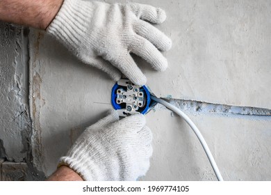 Close-up On The Hands Of A Caucasian Male Electrician Installing A White AC Outlet On A Gray Wall Of A House.Home Repairs During The Pandemic And Stay At Home Period, With Your Own Hands Close-up.