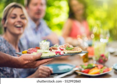 Close-up on hands holding a tray with a preparation of vegetables and parma ham. In the background, friends gathered around the table on the terrace - Powered by Shutterstock