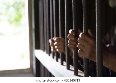 Closeup On Hands Of Man Sitting In Jail. Man Behind Jail Bars On Black Background