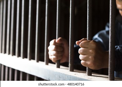 Closeup On Hands Of Man Sitting In Jail. Man Behind Jail Bars On Black Background