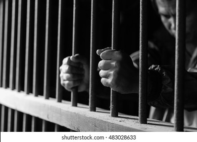 Closeup On Hands Of Man Sitting In Jail. Man Behind Jail Bars With Black And White Style