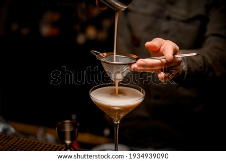 close-up on metal sieve through which male bartender pours frothy espresso martini cocktail into glass