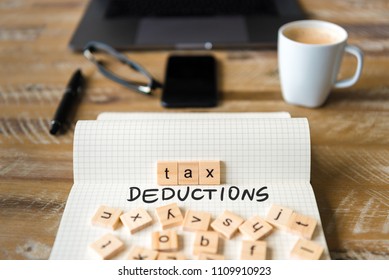 Closeup on notebook over vintage desk surface, front focus on wooden blocks with letters making Tax Deductions text. Business concept image with office tools and coffee cup in background
