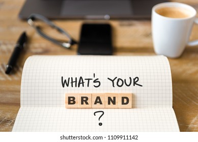 Closeup on notebook over vintage desk surface, front focus on wooden blocks with letters making Whats Your Brand text. Business concept image with office tools and coffee cup in background