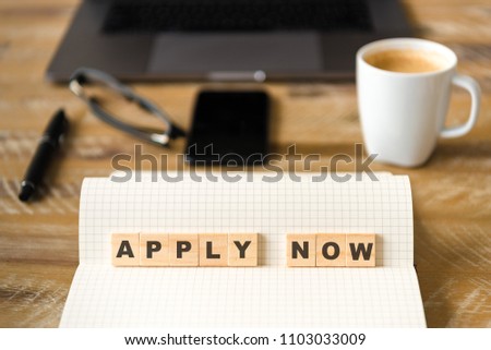 Closeup on notebook over wood table background, focus on wooden blocks with letters making Apply Now text. Concept image. Laptop, glasses, pen and mobile phone in defocused background