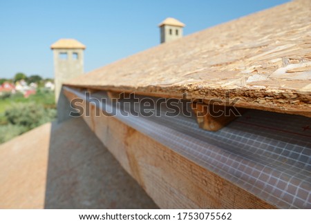 A close-up on plywood board, OSB used for roof sheathing installed on roof beams with blurred roofing construction in the background.