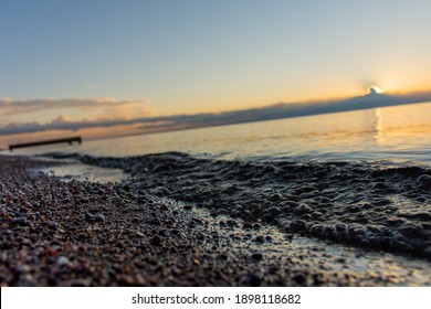 Closeup on sand rocks and small wave with soft focus on wooden dock and rising sun piercing through dark clouds over the horizon and Lake Saint-Jean, Quebec, Canada, on a warm summer morning