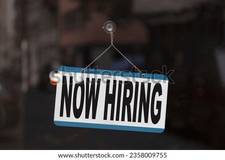 Close-up on a sign in the window of a shop displaying the message: Now hiring.
