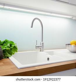 Close-up On Simple, White Kitchen Sink With Silver Faucet, Glass Backsplash, Led Light And Wooden Countertop
