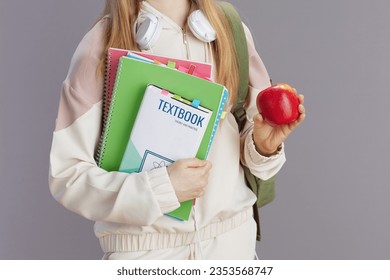 Closeup on teenager girl in beige tracksuit with apple, backpack, workbooks, headphones and textbook isolated on grey background.