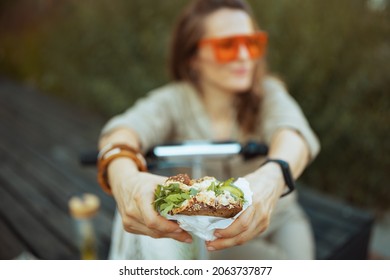 Closeup on woman in sunglasses and overall with scooter and sandwich sitting outdoors in the city.