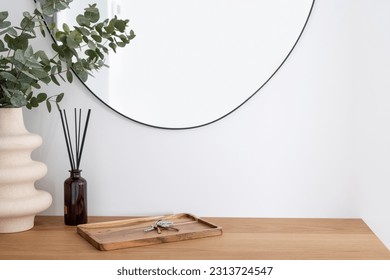 closeup on wooden table surface in hallway at modern apartment, ceramic vase with eucalyptus branch, aroma sticks and keys on bamboo tray against wall with mirror