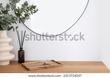 closeup on wooden table surface in hallway at modern apartment, ceramic vase with eucalyptus branch, aroma sticks and keys on bamboo tray against wall with mirror