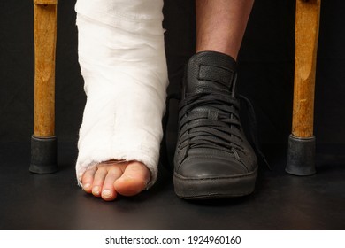 Close-up Of One Broken Leg, In A Cast, The Second Is Shod In A Winter Boot, Next To Crutches On A Black Background.