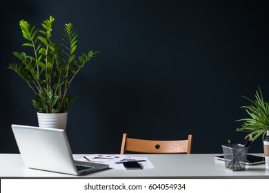 Close-up of open laptop on white table. Nearby is a tablet computer, a smartphone, paper graphics, pencil holder. In the background a dark wall and house plants. Empty workplace.