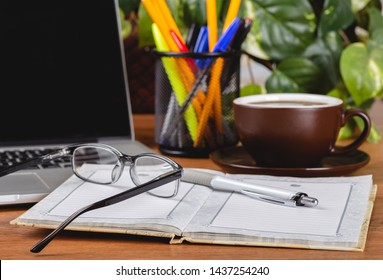 Closeup of an opened notebook, eyeglasses and pen on an office desk with laptop computer ond cup of coffee in background - Powered by Shutterstock