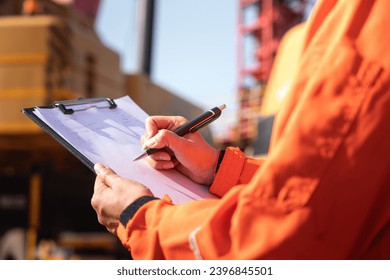 Close-up at the operation supervisor is checking on heavy machine and equipment checklist form with crane truck as blurred background. Industrial safety working in action, selective focus.