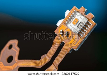 Closeup of optical sensor on electronic printed circuit board and flex ribbon cables on dark blue background. Small orange die in transparent micro chip on PCB of dismantled digital CD-DVD disc drive.
